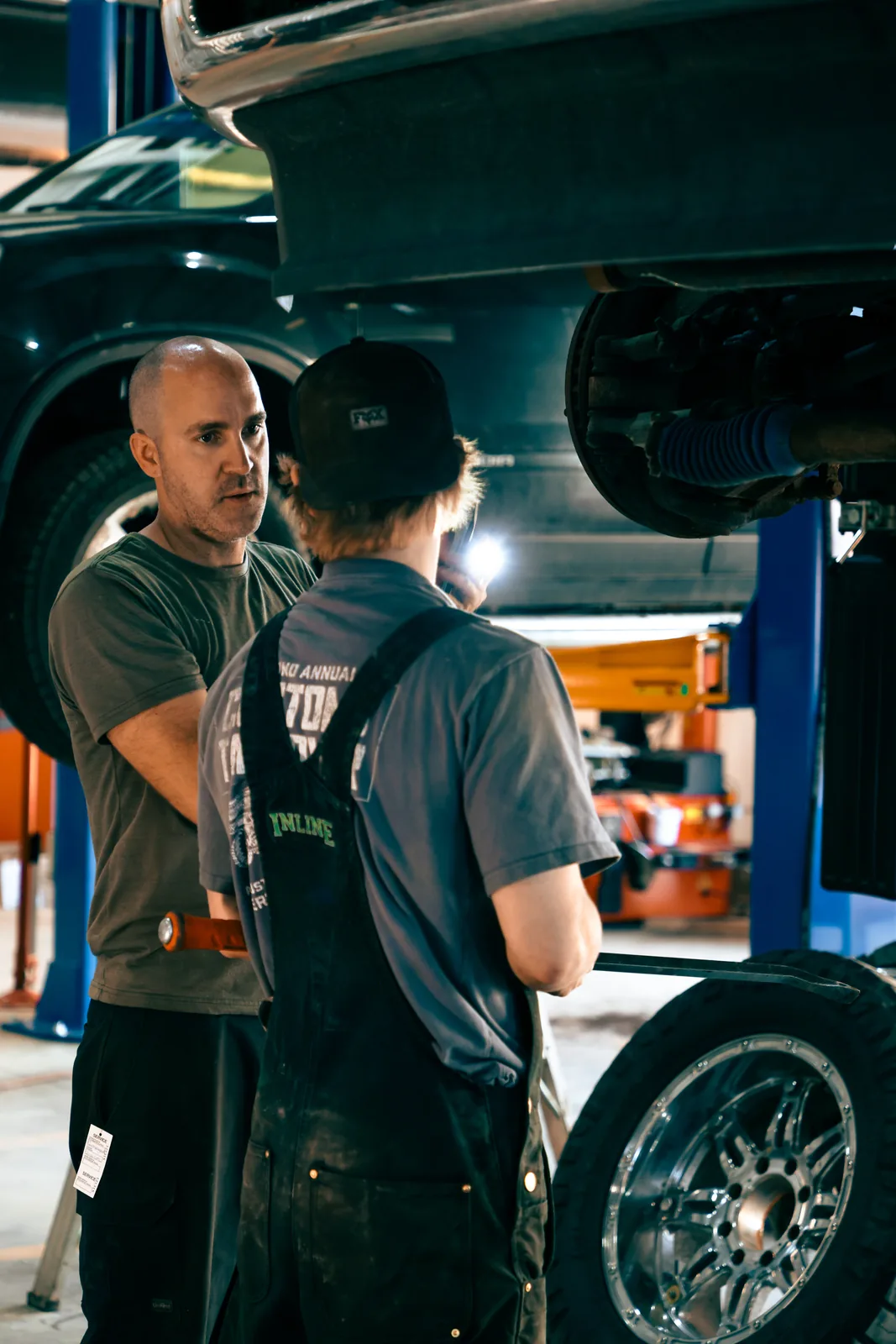 Two men talking beneath a truck raised on a shop hoist