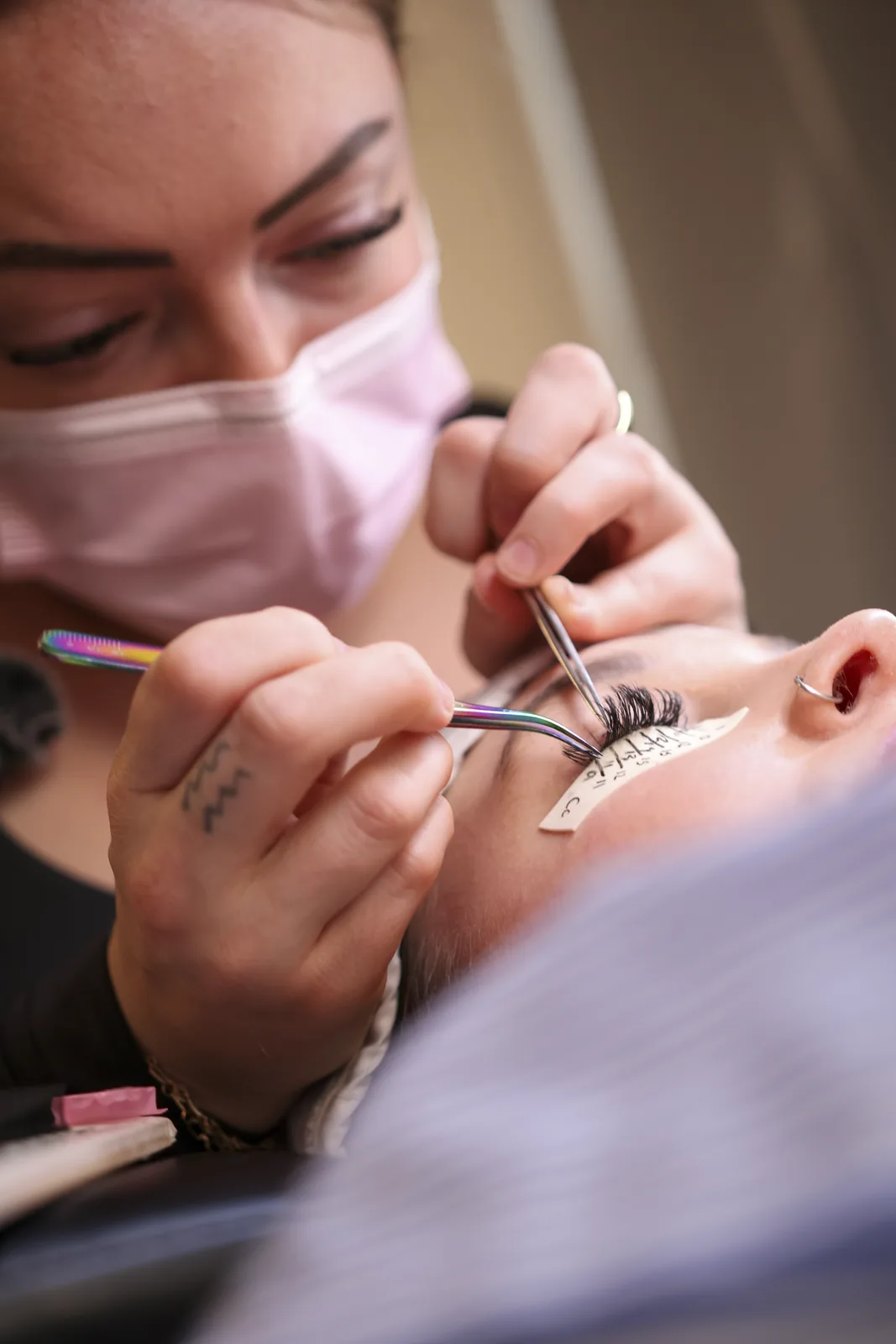 Lash artist in a mask working on a client with fine tweezers